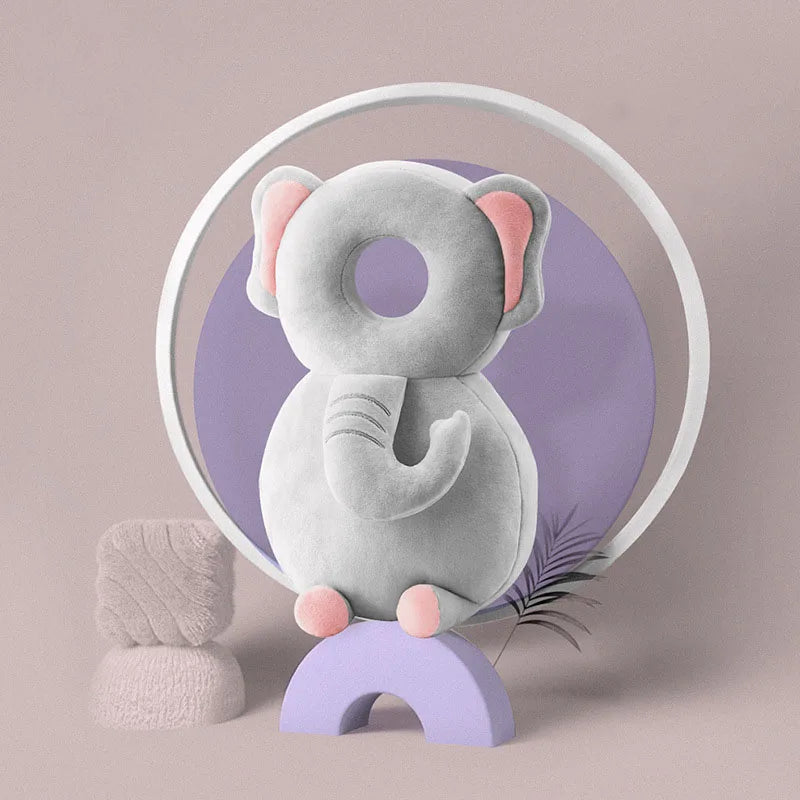 A soft gray elephant-shaped baby head protector pillow with pink ears is displayed inside a white circle against a purple bac