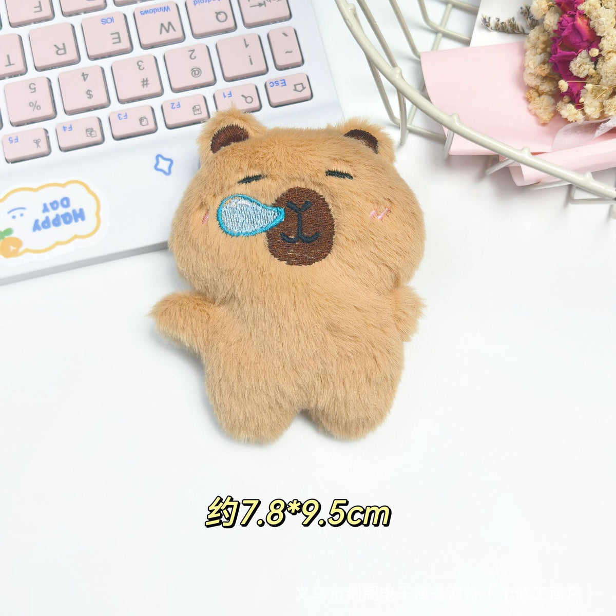 A small, fluffy brown bear plush toy with a light blue nose, resting on a white surface next to a laptop.