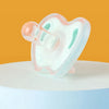 A soft silicone baby pacifier with pink and mint green accents resting on a white surface against an orange background.