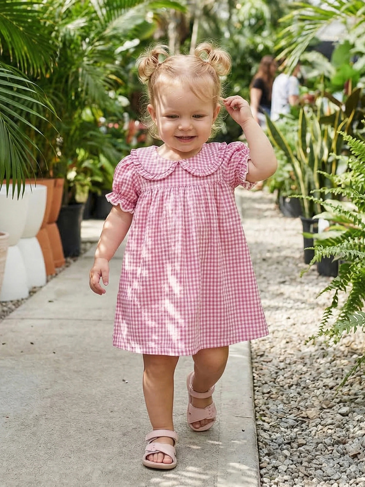 A toddler girl wearing a pink and white gingham A-line dress with a Peter Pan collar walking on a sidewalk in a garden.