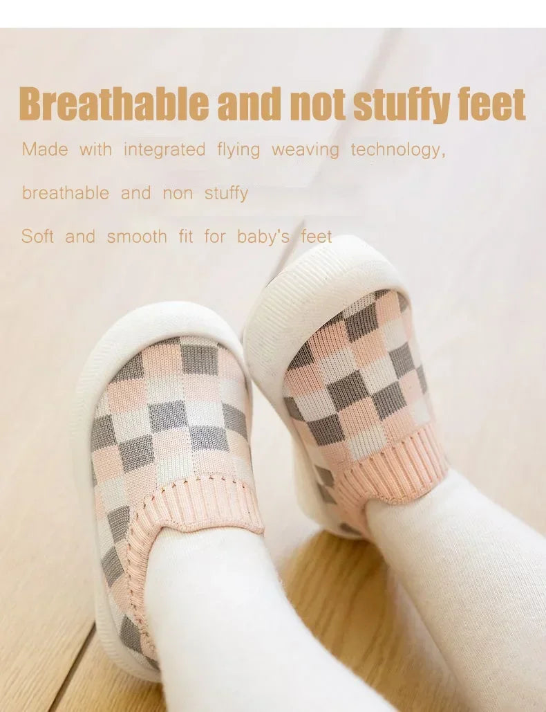 A pair of soft pink and gray plaid slip-on toddler shoes for baby's feet shown on a person's foot.