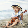 A young boy wearing a straw hat is playing a wooden ukulele toy on a sandy beach.
