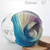 A ball of hand-dyed merino wool yarn in shades of blue, green, and purple is shown on a wooden surface.