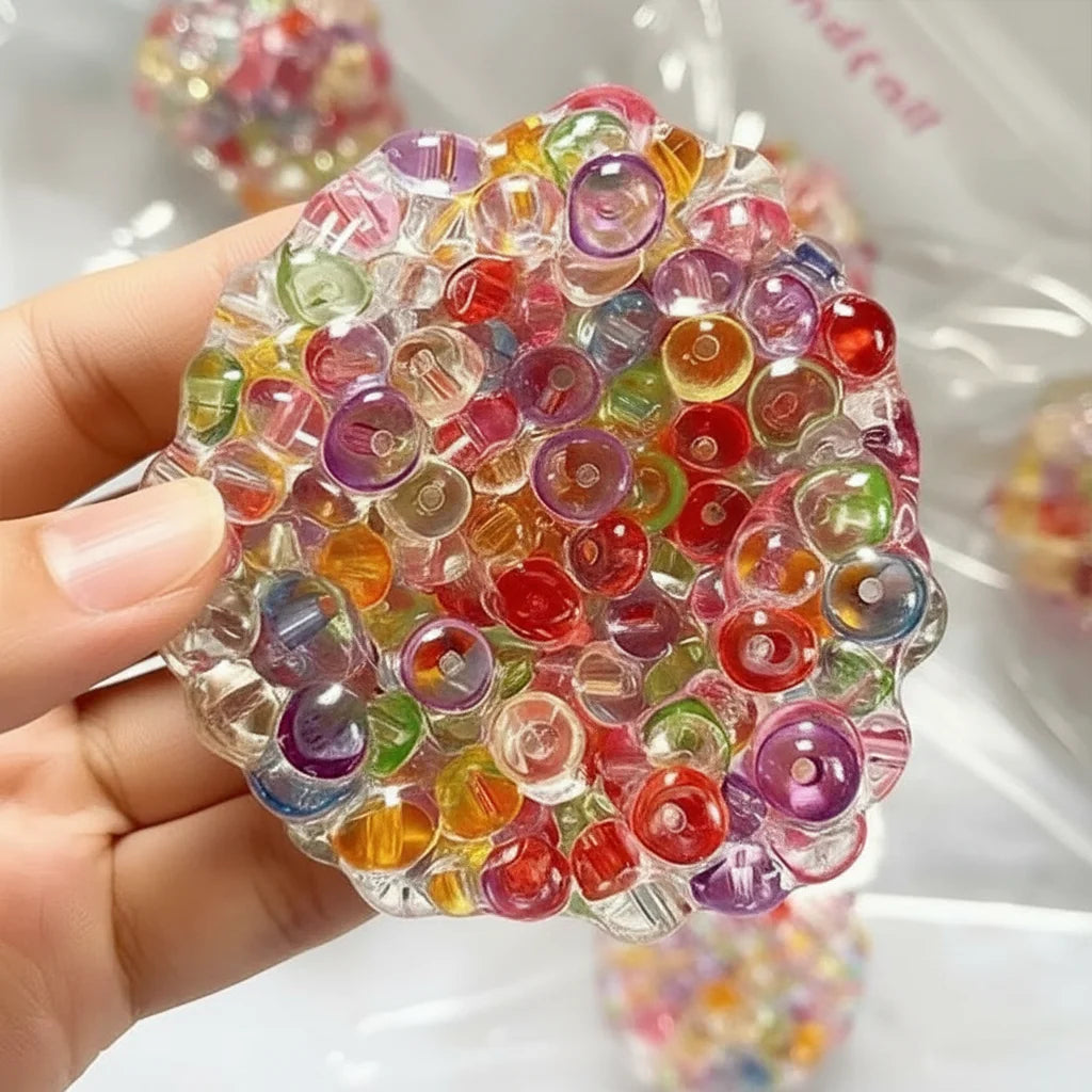 A hand holds a clear, spherical stress ball filled with colorful plastic beads for anxiety relief.