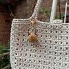A handcrafted crochet animal keychain, resembling a small, round rabbit or chick, is attached to a beige woven bag.