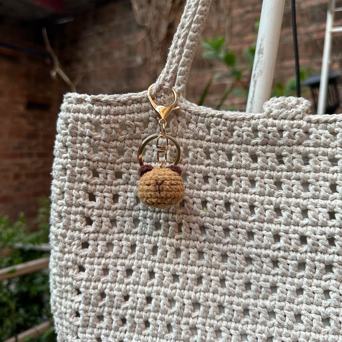 A handcrafted crochet animal keychain, resembling a small, round rabbit or chick, is attached to a beige woven bag.