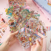 A hand holds up a colorful sticker book featuring 3D cabin scenes and various building elements on pink paper.