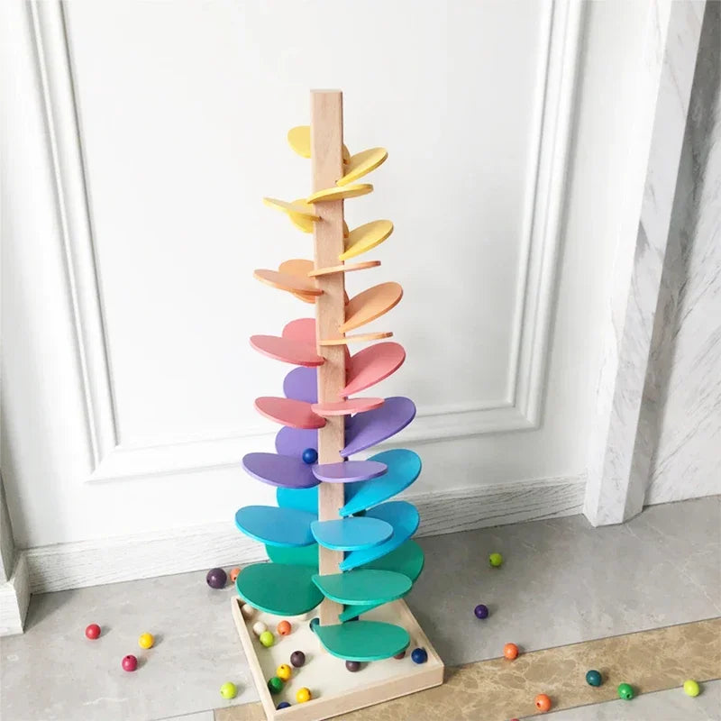 A wooden rainbow ball track toy featuring colorful, layered blocks arranged in a vertical, tree-like structure.