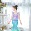 A young girl models a purple and teal mermaid fantasy dress with a ruffled fishtail skirt in a bright, airy room.