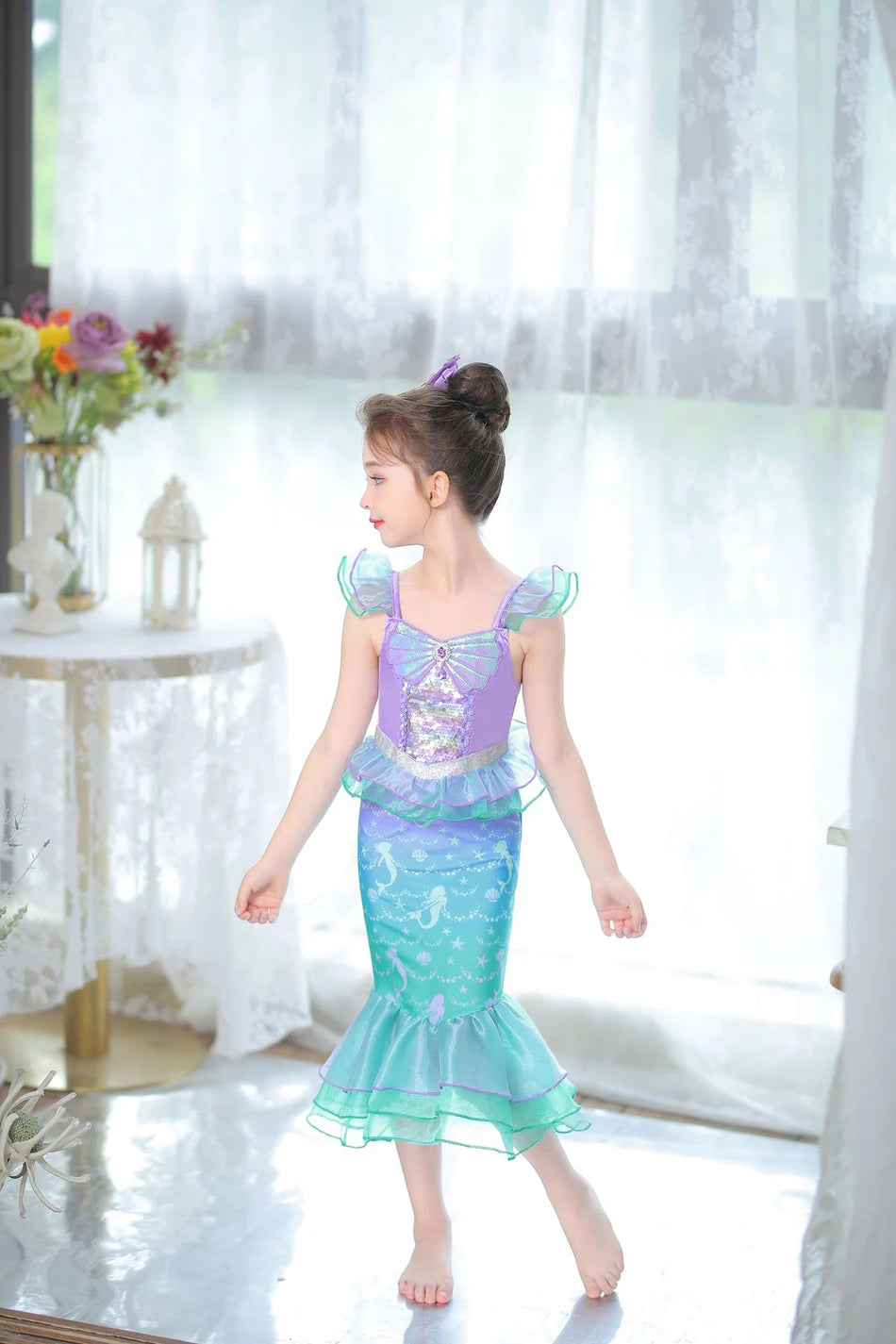A young girl models a purple and teal mermaid fantasy dress with a ruffled fishtail skirt in a bright, airy room.