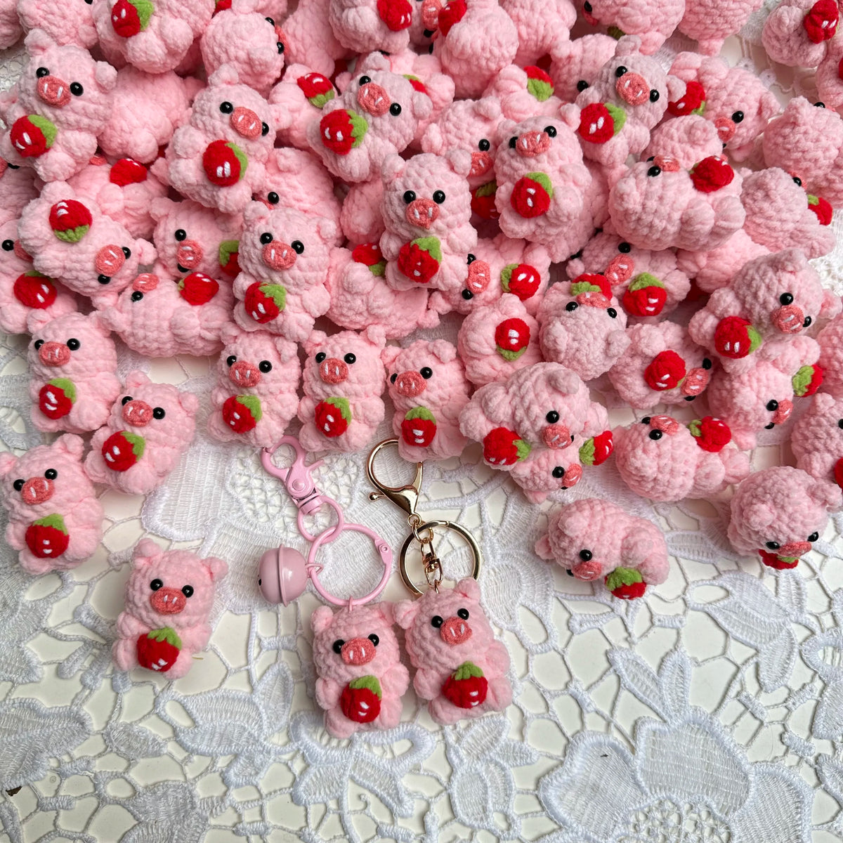A pile of pink crochet mini pig keychains with red accents resting on white lace fabric.