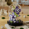 A glass dome terrarium containing purple and pink flowers and moss, resting on a wooden table.