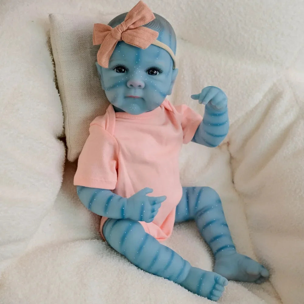 A lifelike reborn baby doll with blue skin and pink clothing is shown resting on a white fluffy surface.