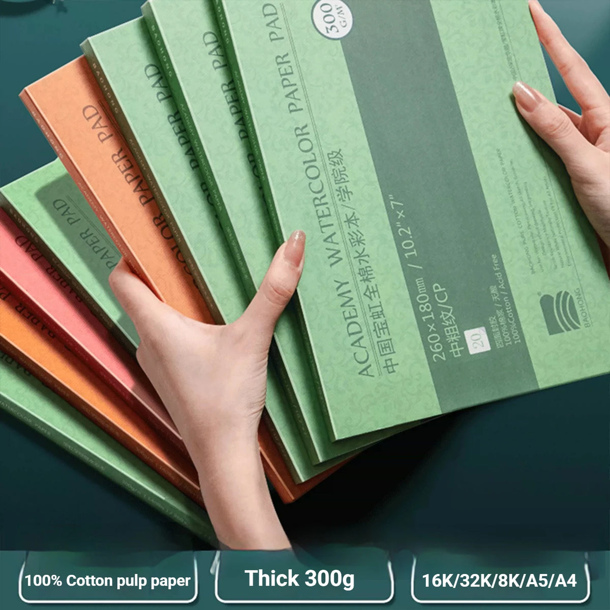 A hand holds a stack of green watercolor paper notebooks with orange and pink covers, showing the thickness and texture.
