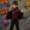 A young boy dressed in a black vampire bat costume with a cape and accessories posing against a Halloween-themed background.