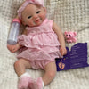 A realistic soft silicone reborn baby doll with a pink outfit and headband sitting on a fluffy white surface.