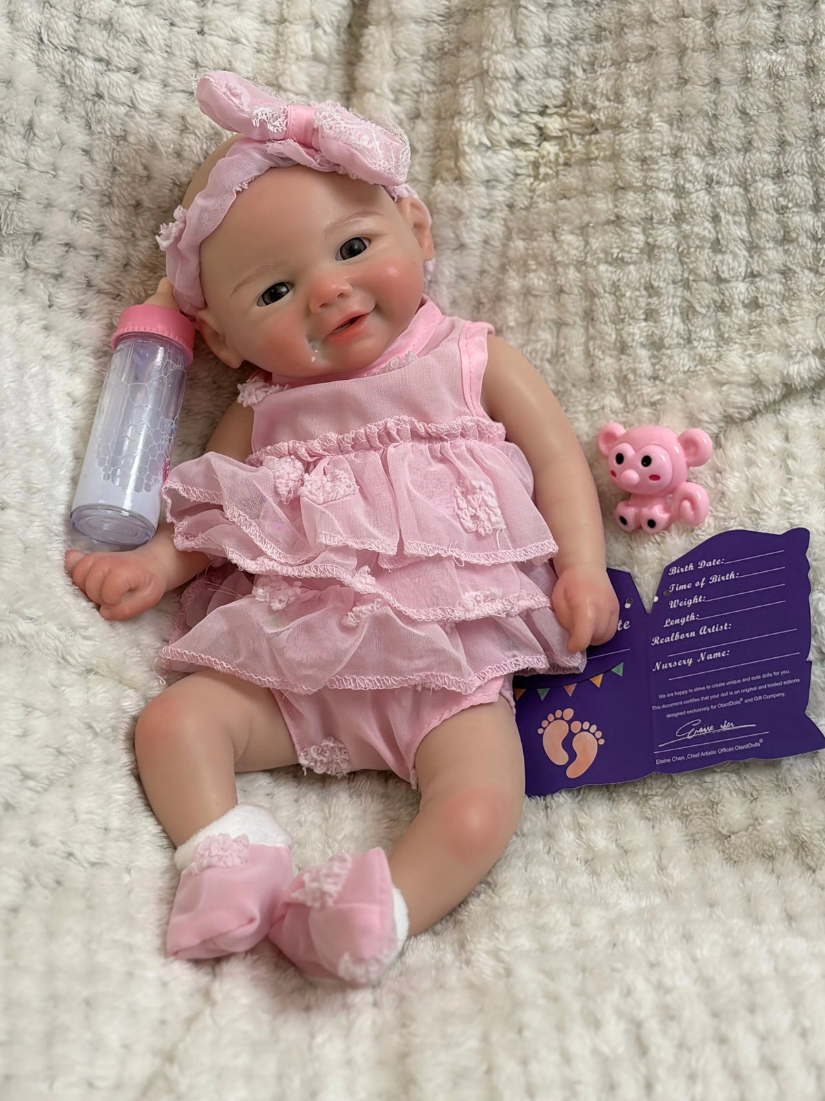 A realistic soft silicone reborn baby doll with a pink outfit and headband sitting on a fluffy white surface.