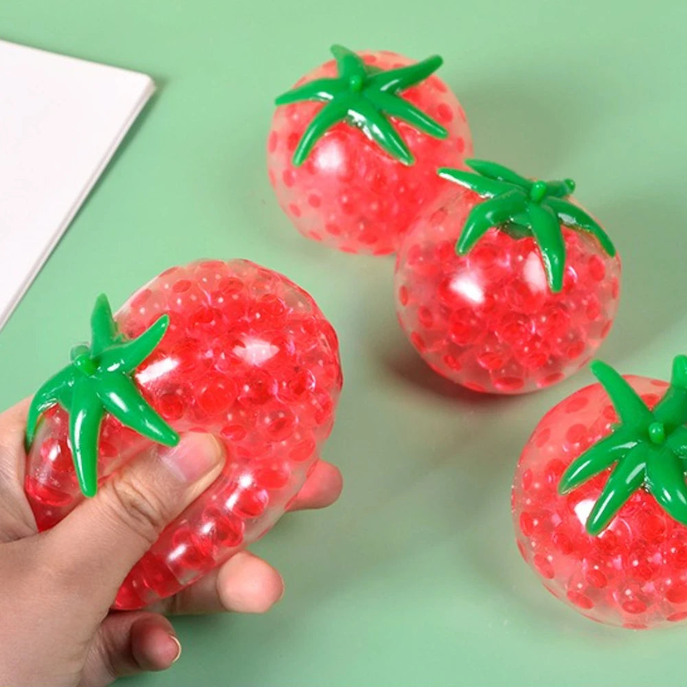 A hand holds a bright red squishy tomato stress relief fidget toy with green leaves against a green background.