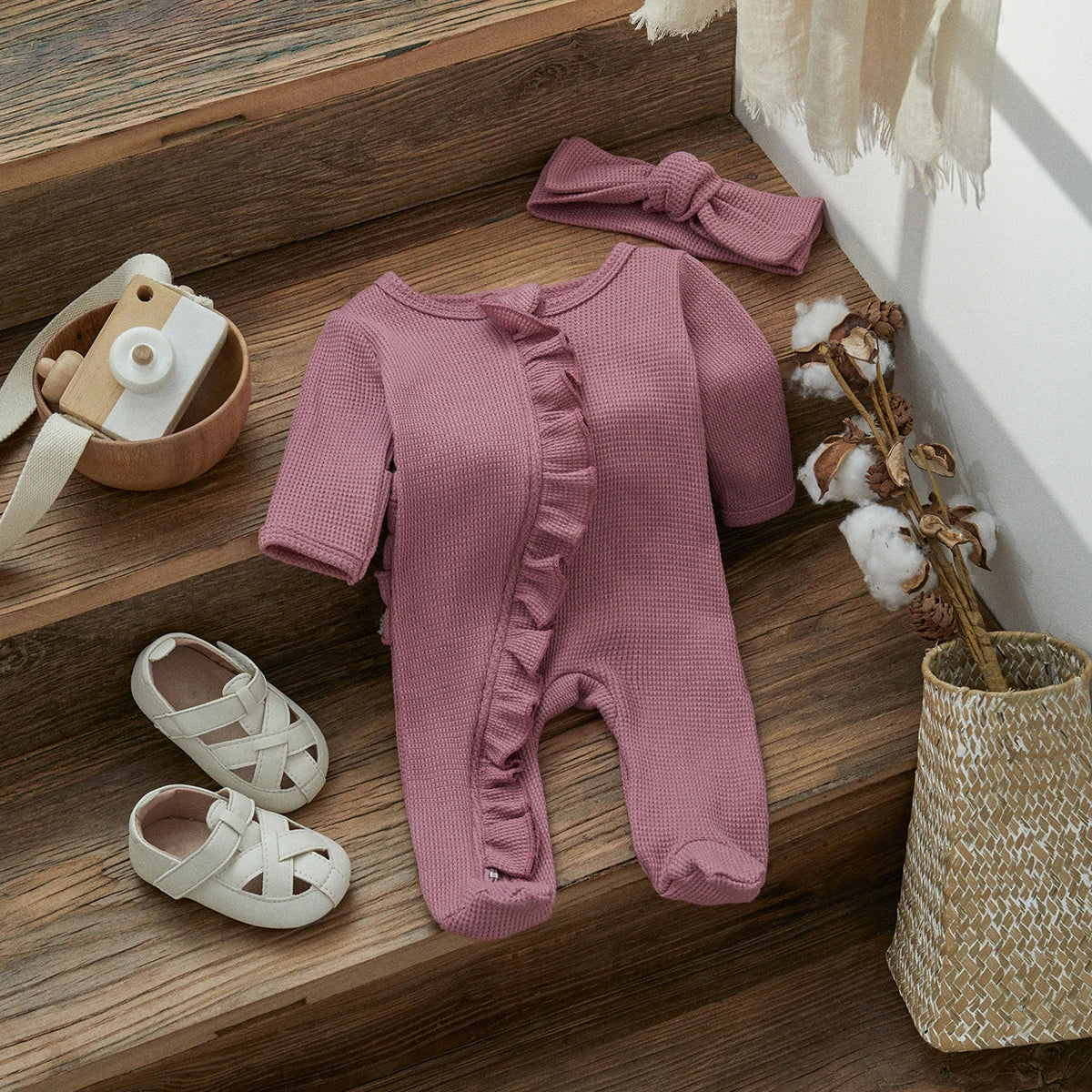 A pink knit ruffle romper for a baby girl displayed on wooden stairs with accessories.