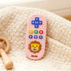 A pink lion-shaped silicone baby teether with remote control buttons rests on a cream-colored textured fabric.