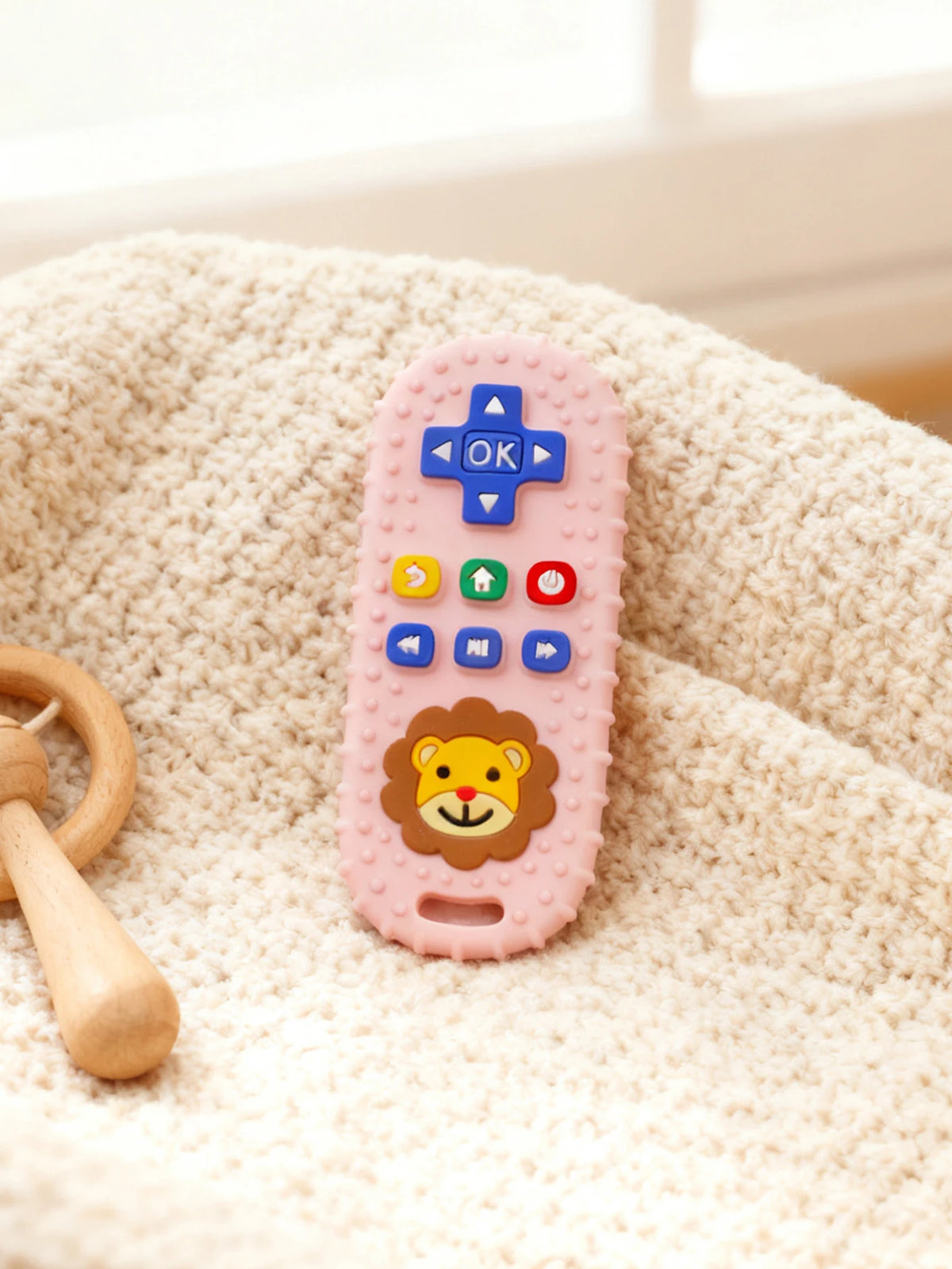 A pink lion-shaped silicone baby teether with remote control buttons rests on a cream-colored textured fabric.