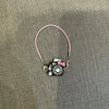 A pink cord necklace with a small black camera charm and a pink bow, used as an accessory for a Labubu doll.