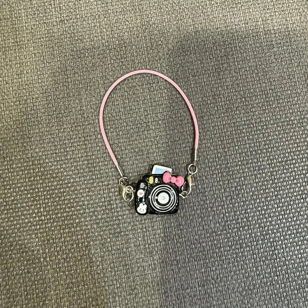 A pink cord necklace with a small black camera charm and a pink bow, used as an accessory for a Labubu doll.