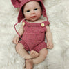 A realistic pink silicone reborn doll wearing a matching knitted rabbit hat and outfit, lying on a fluffy white surface.