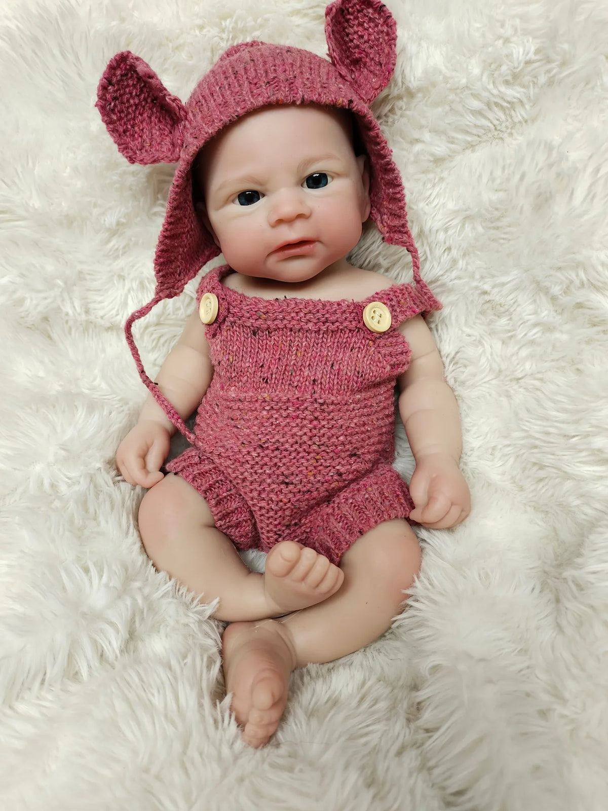 A realistic pink silicone reborn doll wearing a matching knitted rabbit hat and outfit, lying on a fluffy white surface.