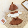 A set of three knitted brown and white doll outfits including a hat, top, and skirt displayed on a woven mat.