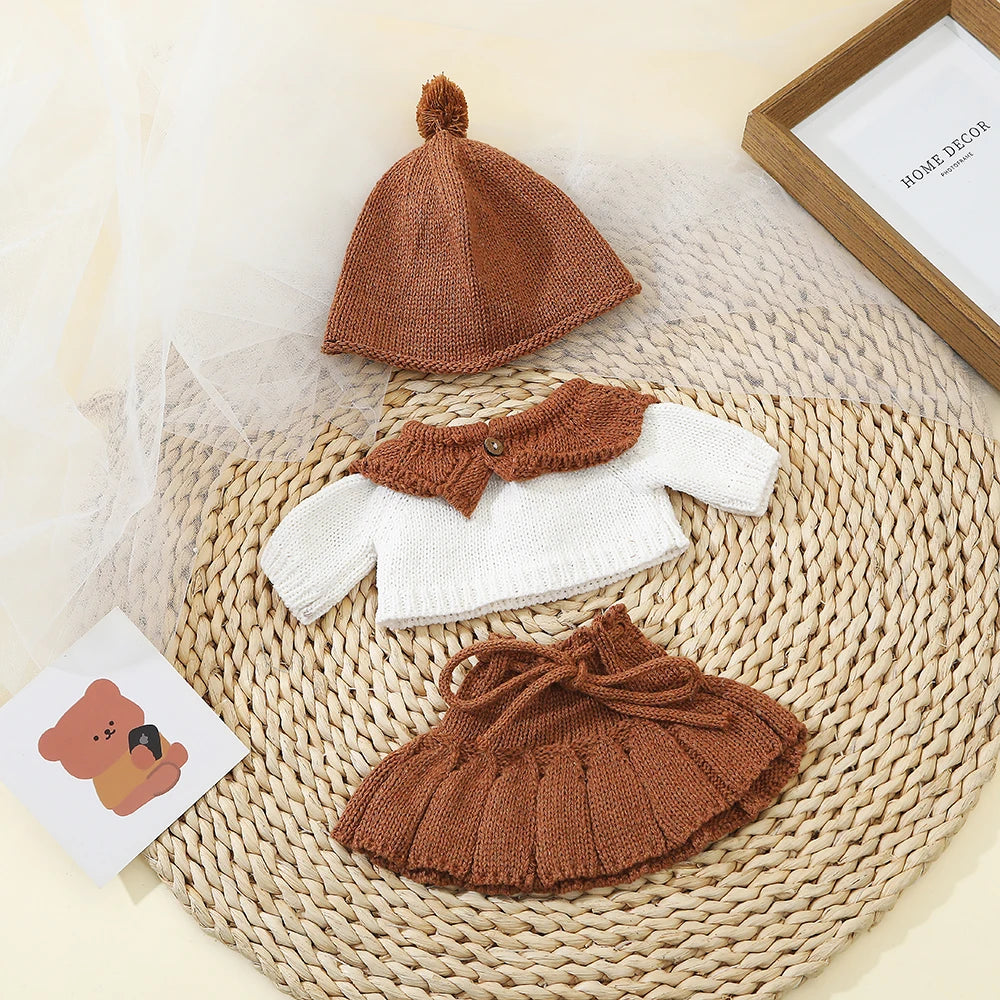 A set of three knitted brown and white doll outfits including a hat, top, and skirt displayed on a woven mat.