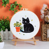 A white embroidery hoop displays a black cat design on a white fabric, set against an orange background.