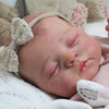 A newborn reborn baby doll with soft cloth body is sleeping peacefully on a white blanket.