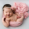 A newborn baby is sleeping wrapped in a pink knit photography wrap with a pearl headband.