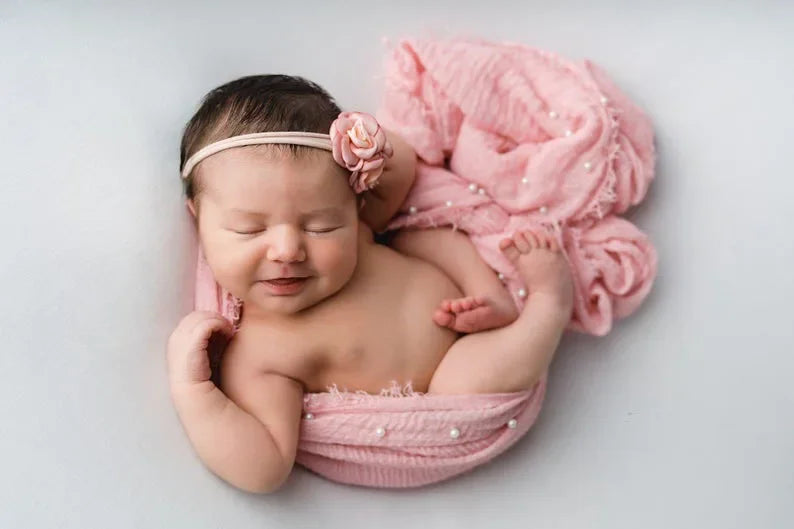 A newborn baby is sleeping wrapped in a pink knit photography wrap with a pearl headband.