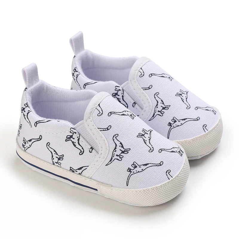 A pair of white baby first walker sneakers with a black dinosaur print on the canvas upper.