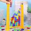 A yellow plastic puzzle frame filled with colorful translucent building blocks and a hand inserting a purple block.