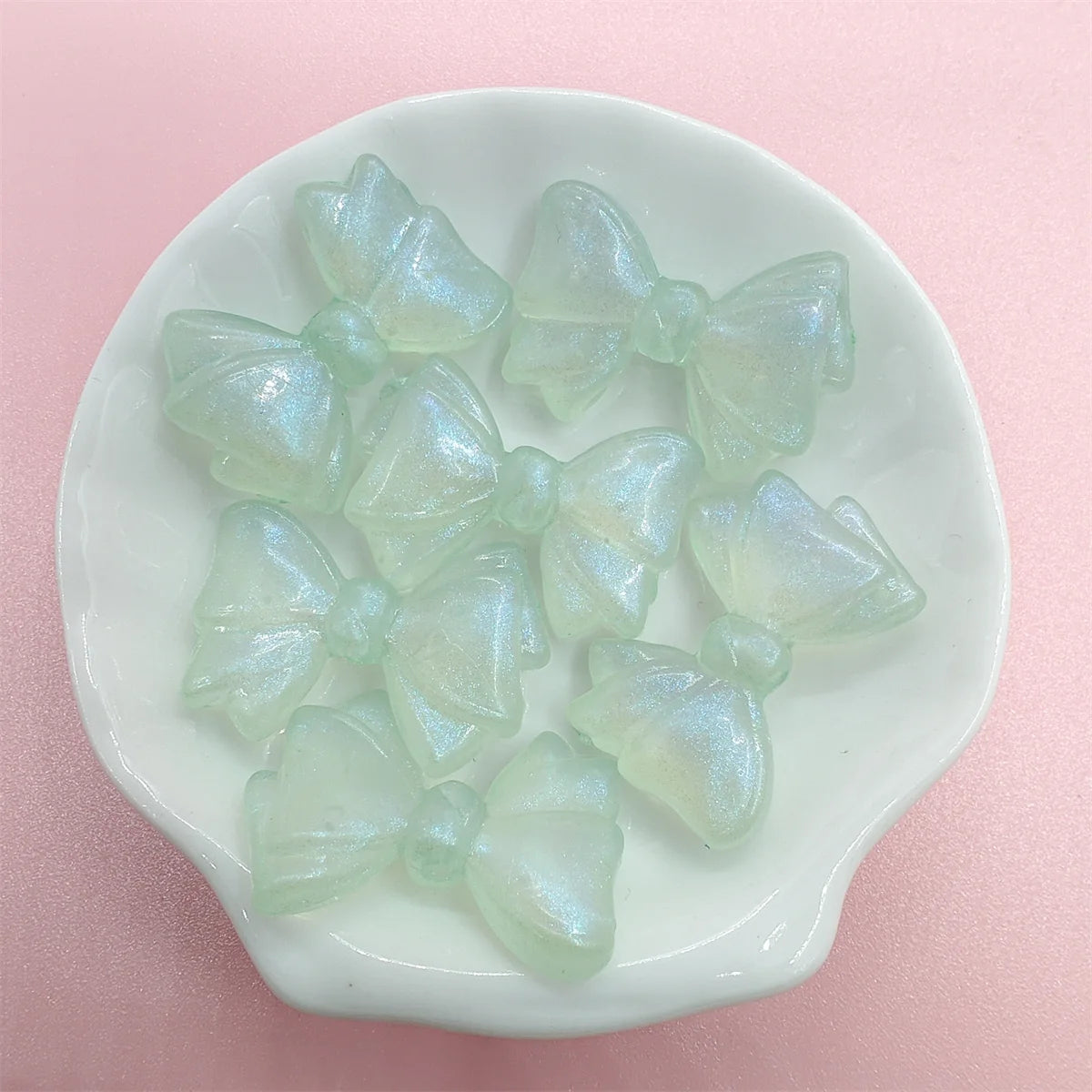 A collection of translucent, pale green shimmer mermaid bowknot beads arranged on a white dish.