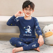 A young boy wearing a navy blue cotton pajama set with an astronaut print, sitting on the floor.