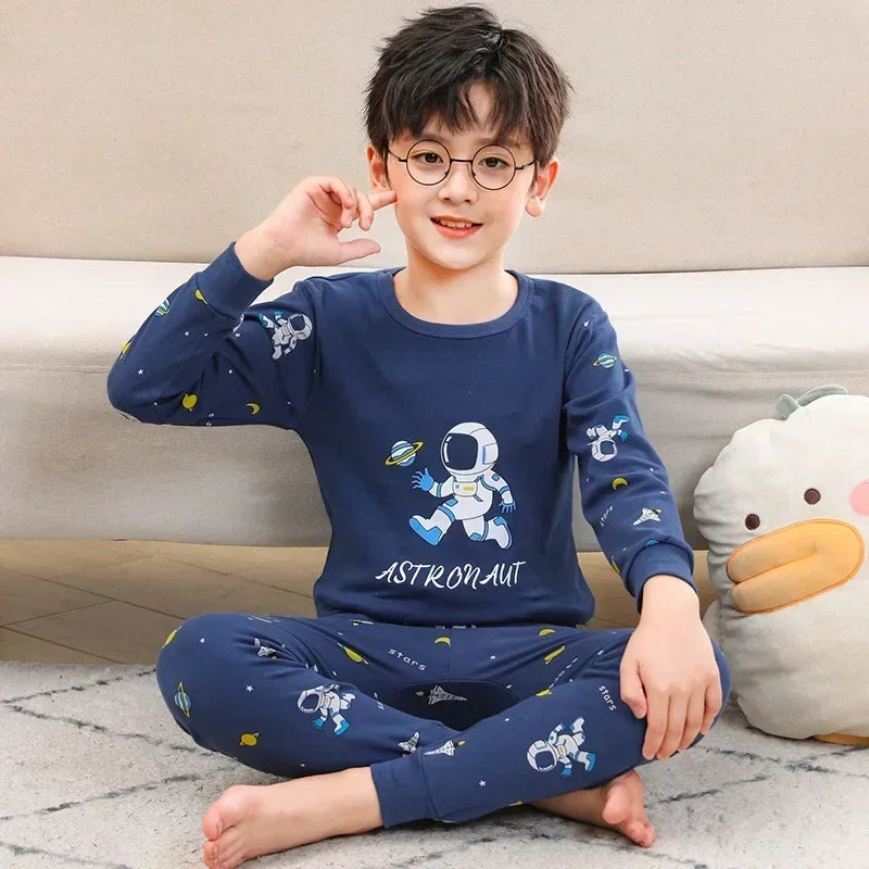 A young boy wearing a navy blue cotton pajama set with an astronaut print, sitting on the floor.