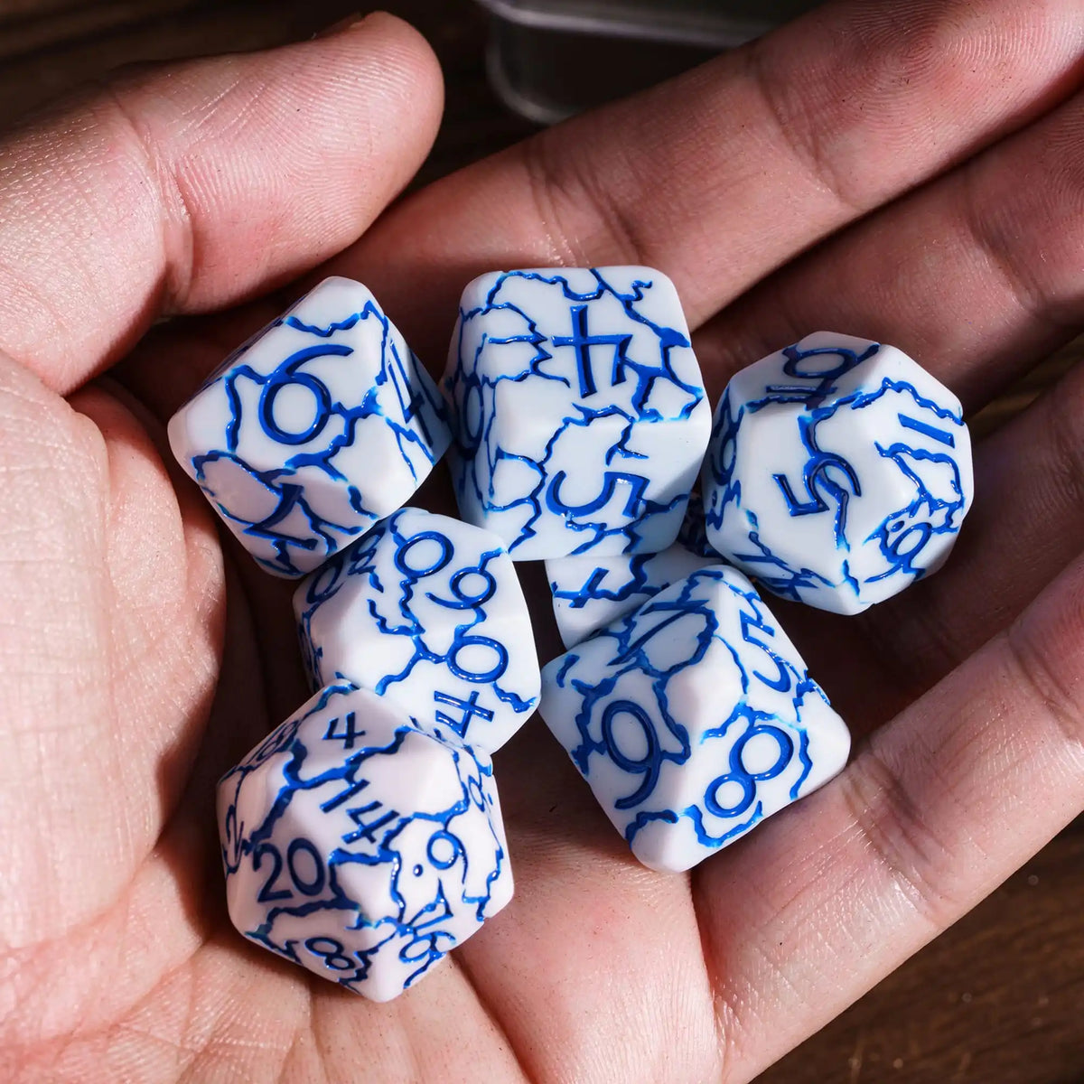A hand holds seven frosted blue and white polyhedral dice on a wooden surface.