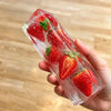 A hand holds a clear ice cube shaped like a strawberry with green leaves inside.