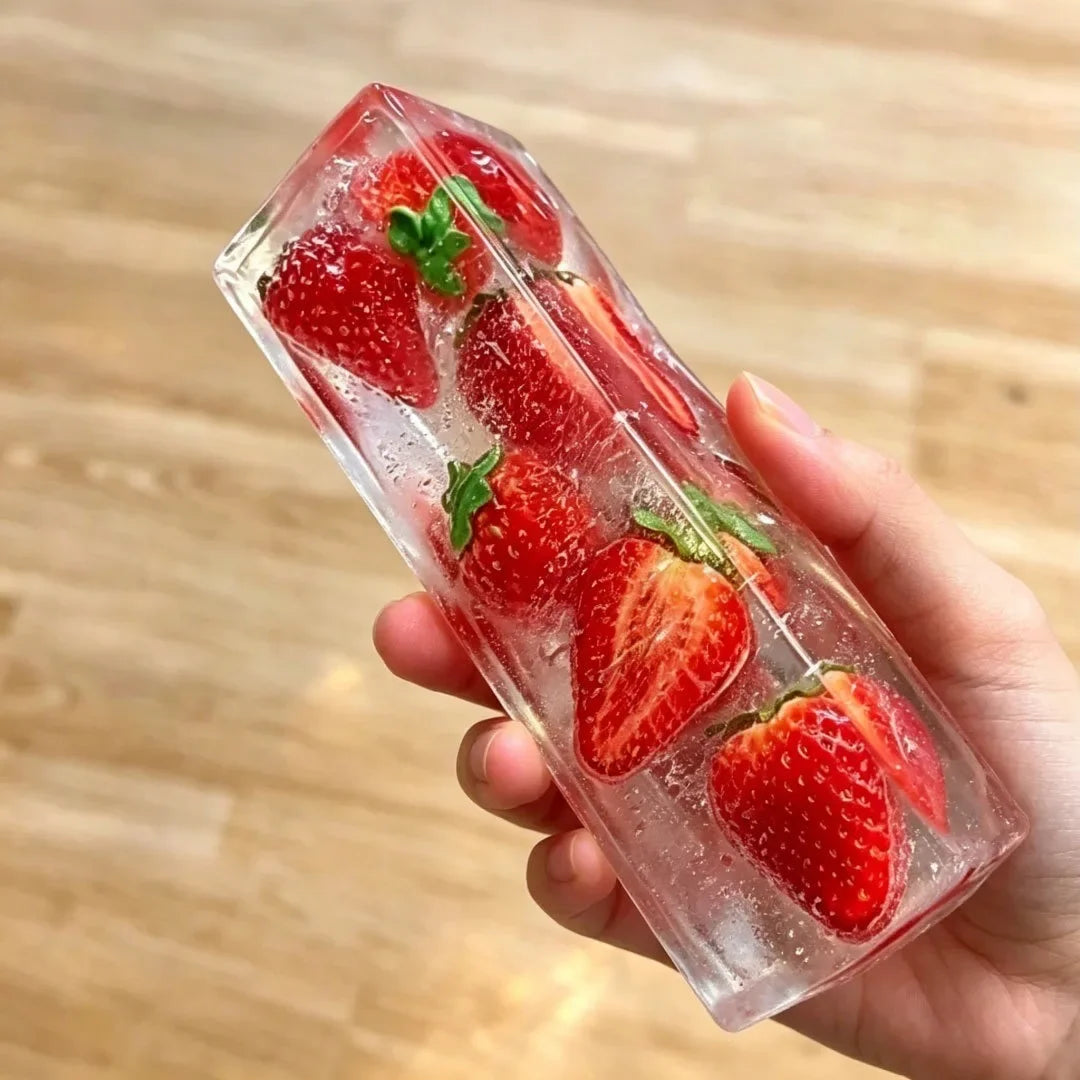 A hand holds a clear ice cube shaped like a strawberry with green leaves inside.