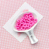 A white spoon filled with pink acrylic beads on a pink grid background.