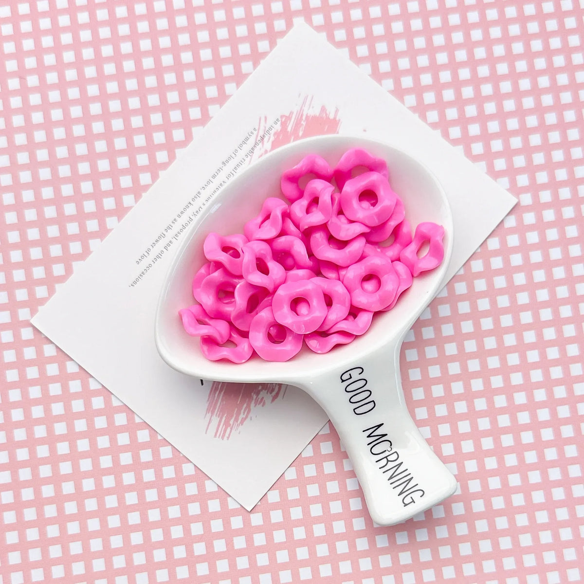 A white spoon filled with pink acrylic beads on a pink grid background.