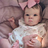 A close-up photo of a lifelike 18-inch reborn baby doll with hand-painted details wearing a floral dress and pink bow, restin