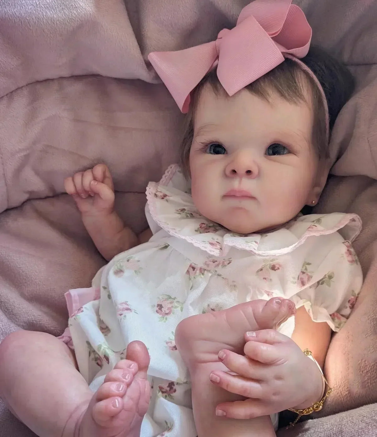 A close-up photo of a lifelike 18-inch reborn baby doll with hand-painted details wearing a floral dress and pink bow, restin