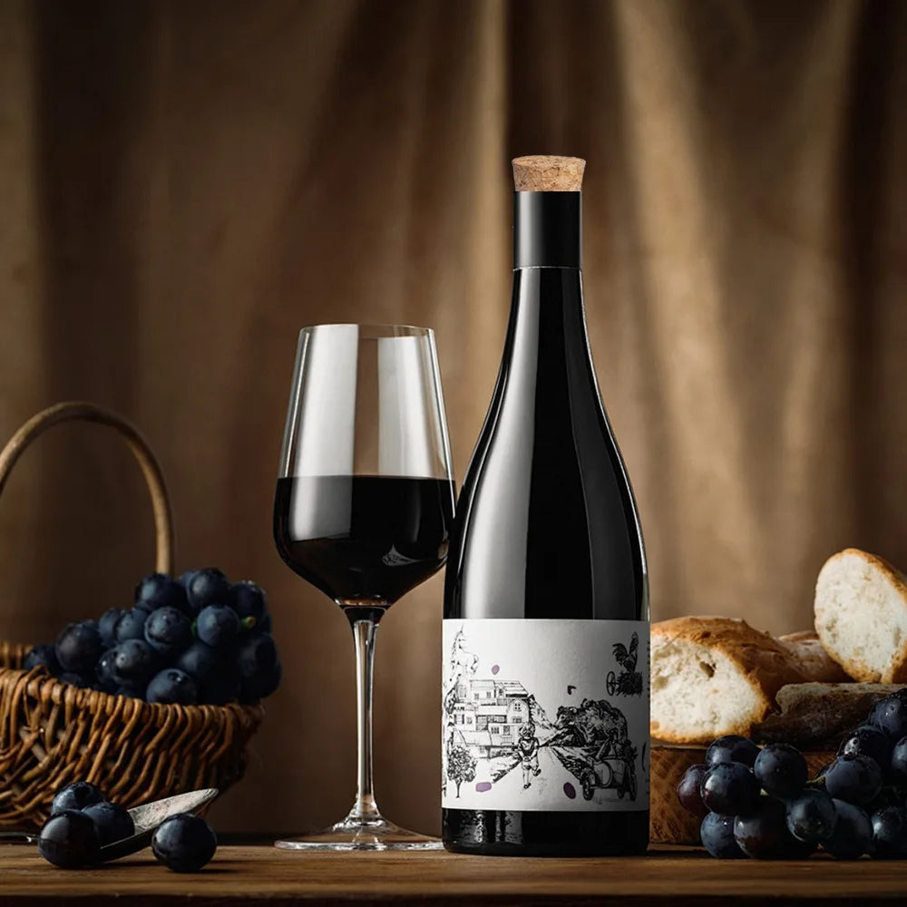 A dark glass bottle of red wine stands next to a wine glass, grapes, and bread on a wooden surface.