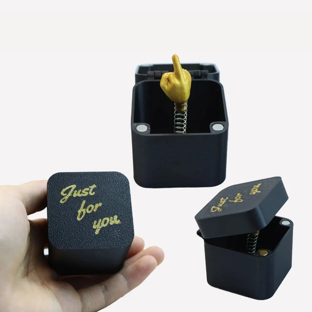 A black pop-up gift box with a yellow middle finger figure is shown being held in a hand.