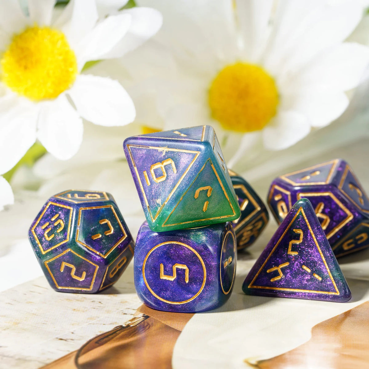 A set of seven purple and green polyhedral dice resting on a wooden surface with white flowers in the background.